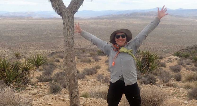 A person smiles and raises their hands in the air while standing in a vast desert landscape